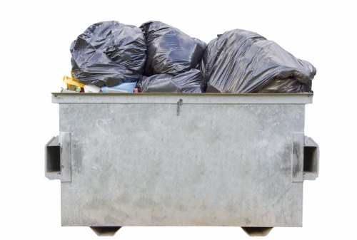 A large metal skip filled with multiple black refuse bags, stacked and slightly bulging, situated outdoors on a paved surface. The skip is rectangular with a dull, light grey finish and has two cut-out handles on each side. In the background, the environment appears to be an industrial or residential area, with no other objects or structures visible around the skip. The black rubbish bags show some creases and folds, indicating they are filled with debris, and they occupy most of the space within the skip. The scene suggests a recent rubbish collection or clearance, typical of garden or property waste removal services in the Herne Hill area, provided by Garden Clearance Herne Hill, focusing on general rubbish disposal or clearance tasks.