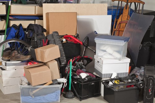 A collection of various items gathered for rubbish removal, including several cardboard boxes in different sizes, some sealed and others open revealing packed contents, stacked on top of each other. There is a clear plastic container with a blue lid, filled with bundled paper, positioned on the right side. A black toolbox or storage bin sits near the centre containing tools or miscellaneous objects, with a green broom handle partially visible behind it. A red and black garden trimmer or hedge cutter leans against the boxes, with its power cord trailing across the scene. To the left, there is a large grey plastic laundry basket filled with fabric or soft materials, and some black backpacks or bags are also present. The background shows a variety of furniture, including a wooden chair with vertical slats and blue support, as well as a grey mesh outdoor chair or recliner, suggesting an indoor or outdoor storage or clearance setting in Herne Hill. The scene depicts a typical arrangement of household or garden waste awaiting collection by Garden Clearance Herne Hill for rubbish disposal services, with natural lighting highlighting the mixed textures and colors of the items.