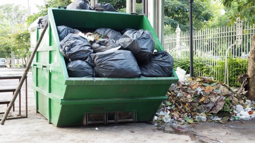 A large green metal skip filled with multiple black plastic rubbish bags, situated outdoors on a paved area next to a garden fence and trees, with some loose waste and discarded materials scattered nearby on the ground. The skip appears to be used for garden or general waste collection, with its surroundings suggesting an urban environment typical of Herne Hill. The scene captures the practical aspect of rubbish removal services provided by Garden Clearance Herne Hill, emphasizing the handling of bulky waste in residential or community settings, with natural daylight illuminating the scene and highlighting the textures of the bags, metal, and nearby environmental features.